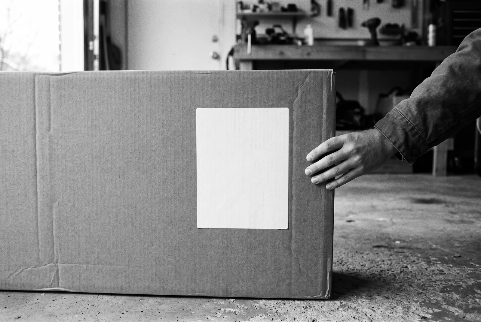 Render of a hand steadying a large flat cardboard carton in a garage, with a blank white label on its side.