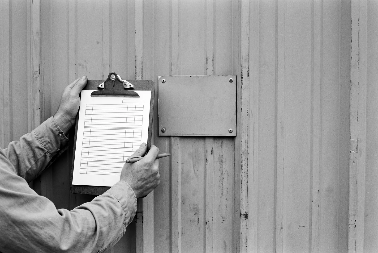 Render of hands holding a clipboard with a blank lined form beside a small blank metal plate riveted to ribbed metal siding.