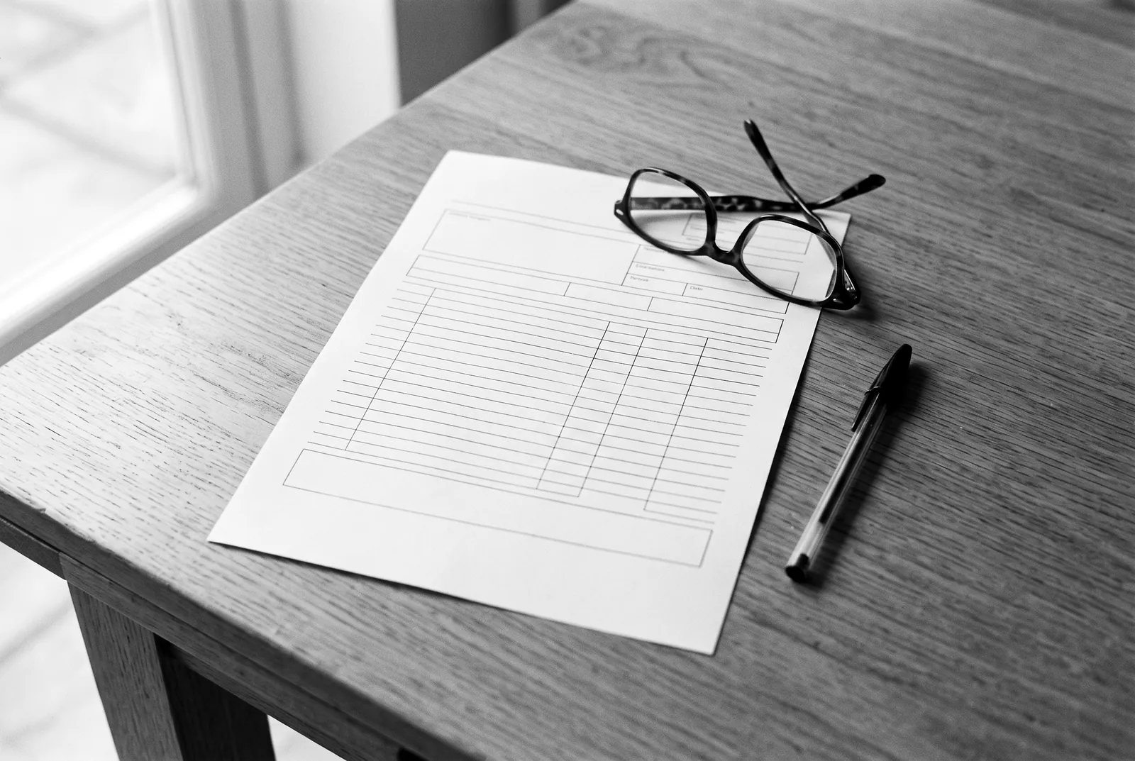Render of a printed sheet of empty ruled boxes on a wooden table with reading glasses and a pen.