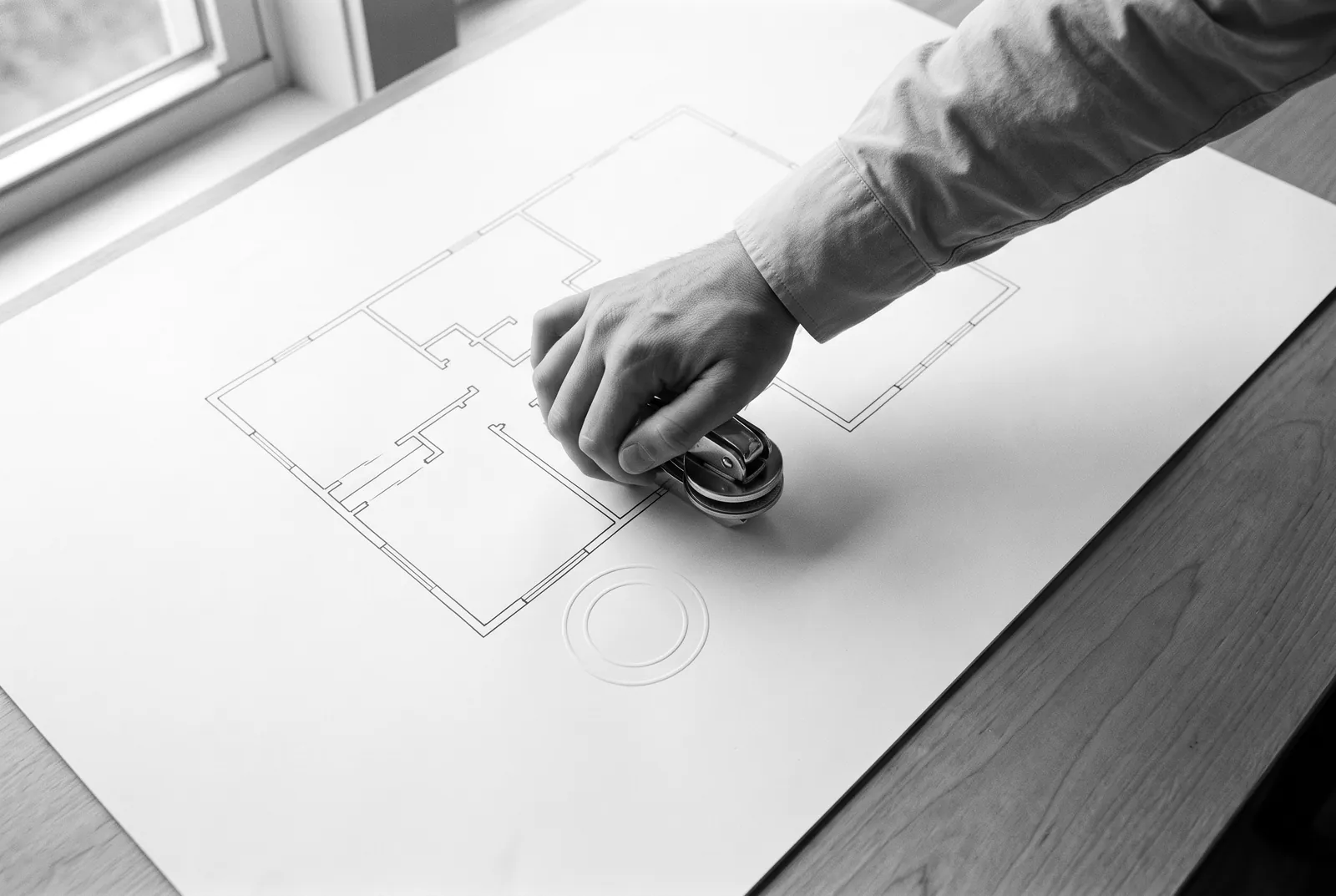Render of a hand pressing a round seal embosser onto a large sheet with a floor plan drawn in thin lines, beside a blank raised seal.