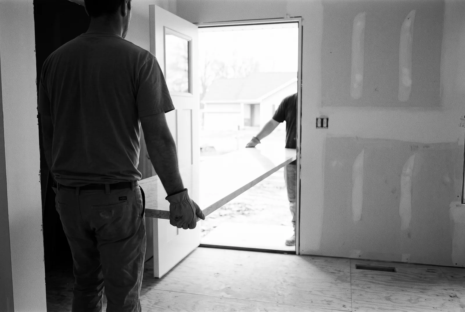 Render of two workers in gloves carrying a pale stone countertop slab through the open front door of a house.