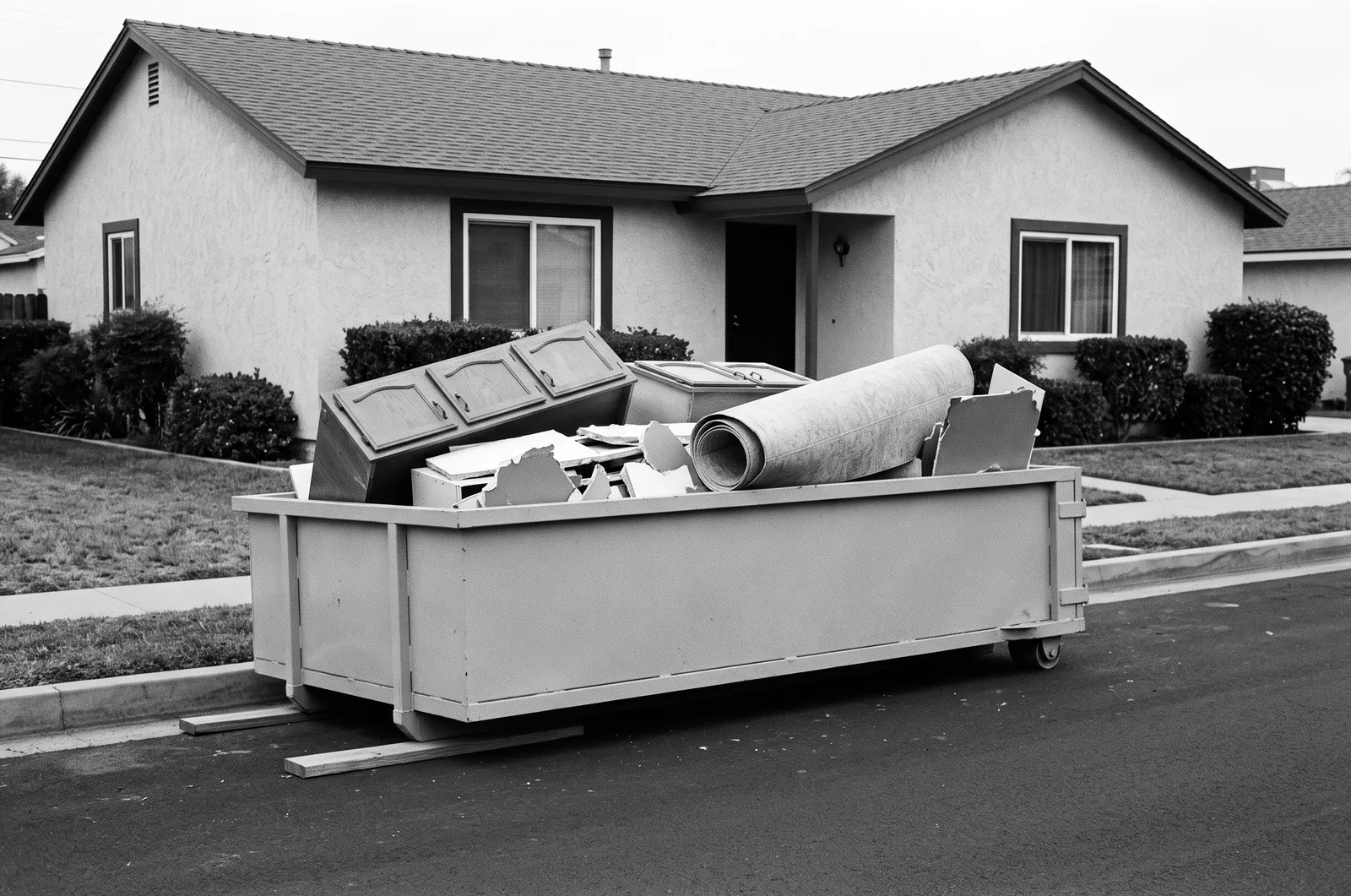 Render of a plain steel roll off bin on wooden boards at a street curb, holding old cabinets, broken drywall and a roll of sheet flooring.