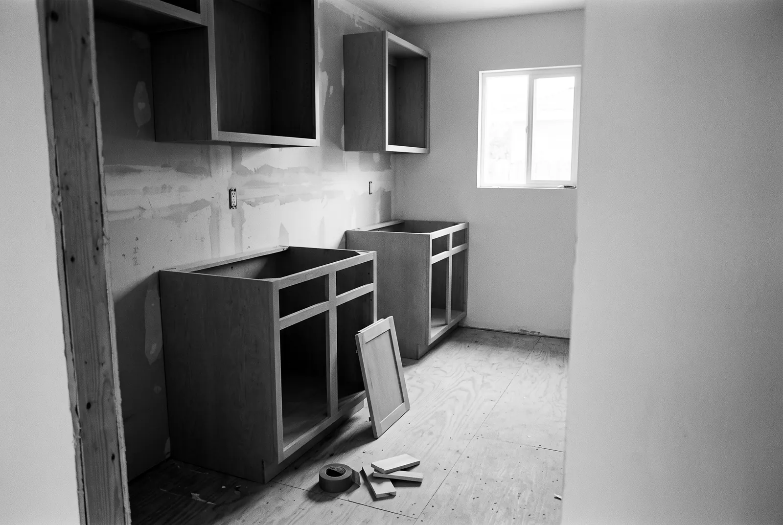 Render of a half finished galley kitchen with doorless cabinet boxes, patched drywall and a loose cabinet door leaning on the plywood floor.