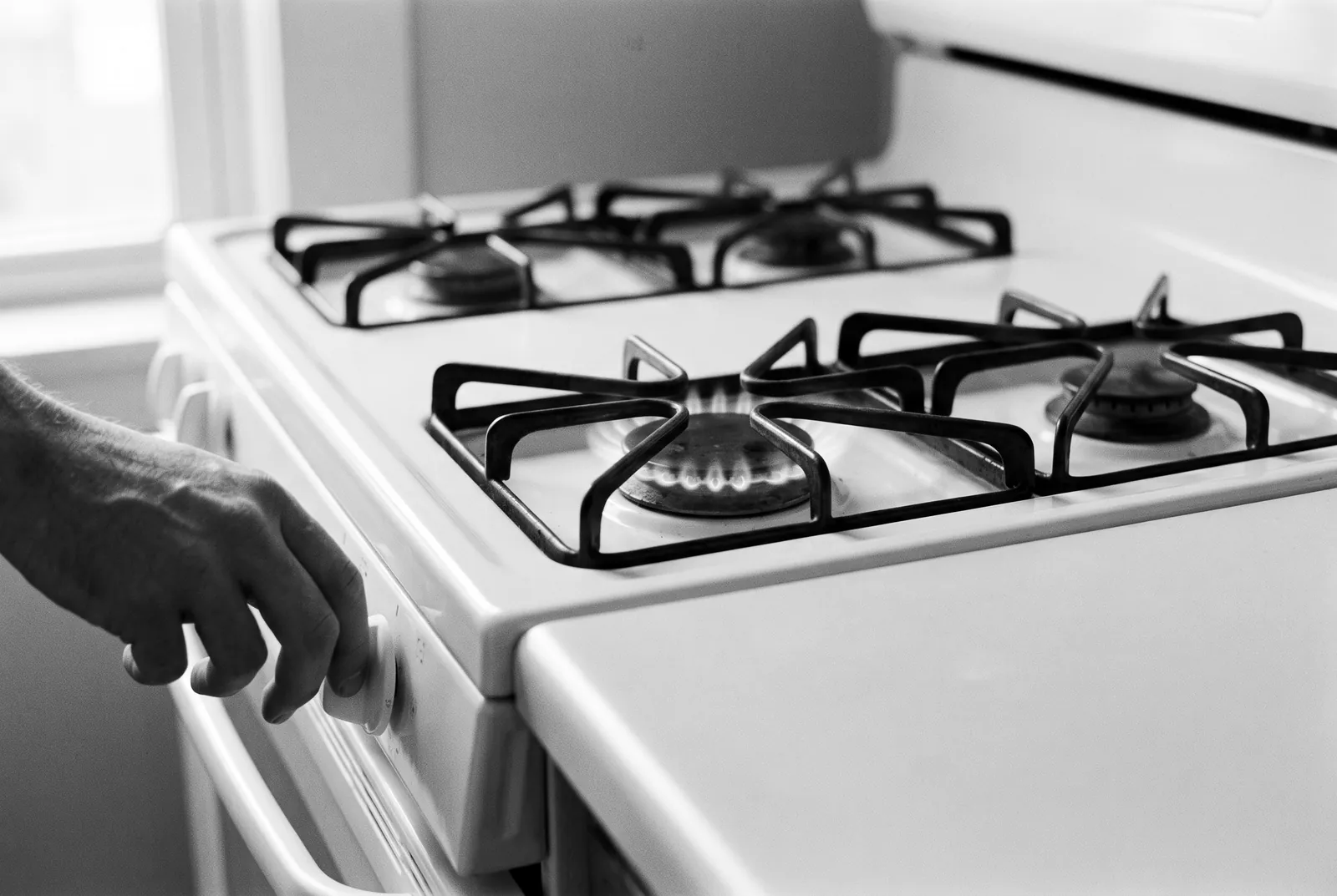 Render of a hand turning the front knob of a white gas range, one burner lit under an empty grate.
