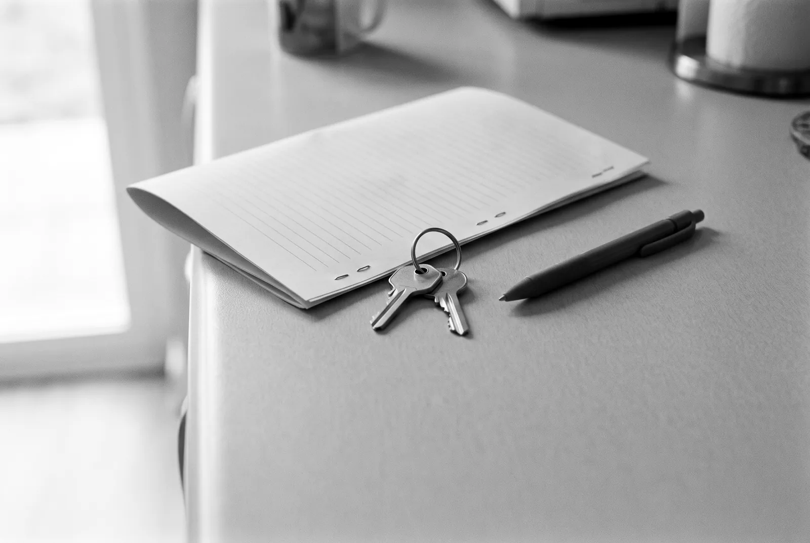 Render of two house keys on a ring, a stapled folded document with blank ruled pages and a pen on a counter.
