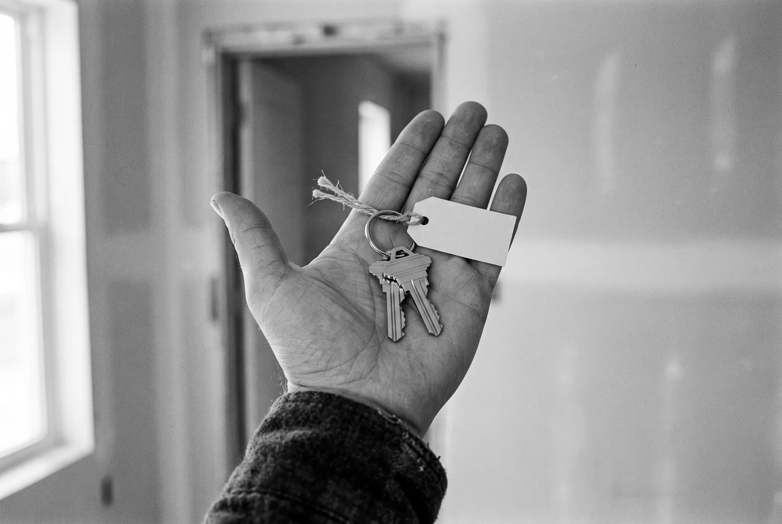 Render of an open hand holding two new house keys on a ring with a blank paper tag, a bare doorway behind.
