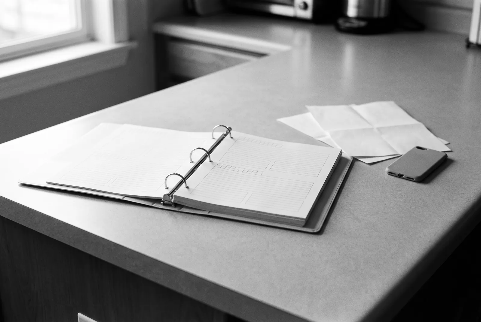 Render of an open binder with blank ruled pages on a kitchen counter, with folded sheets and a phone beside it.