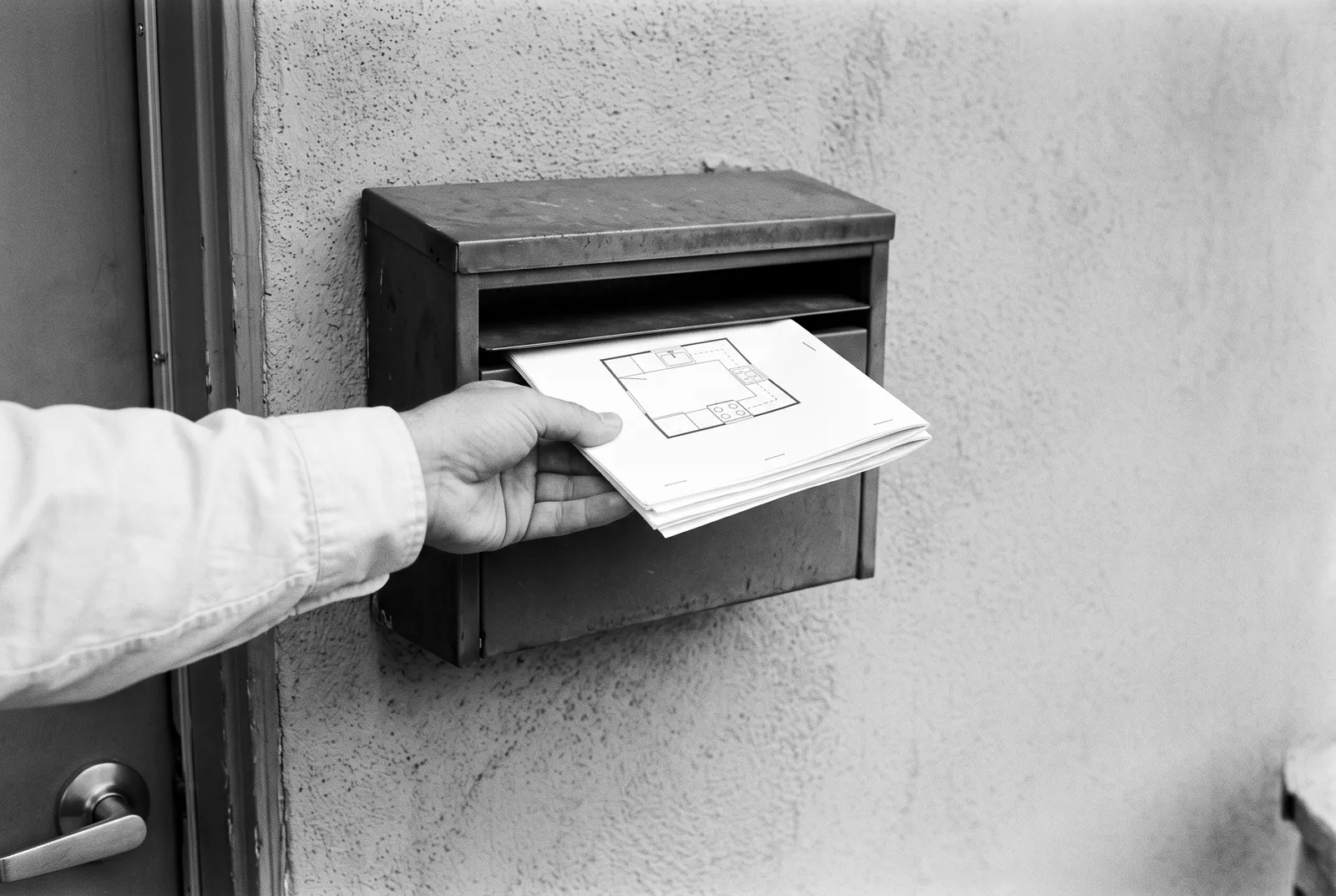 Render of a hand sliding a stapled packet with a simple kitchen floor plan drawing into a metal drop box on a stucco wall.