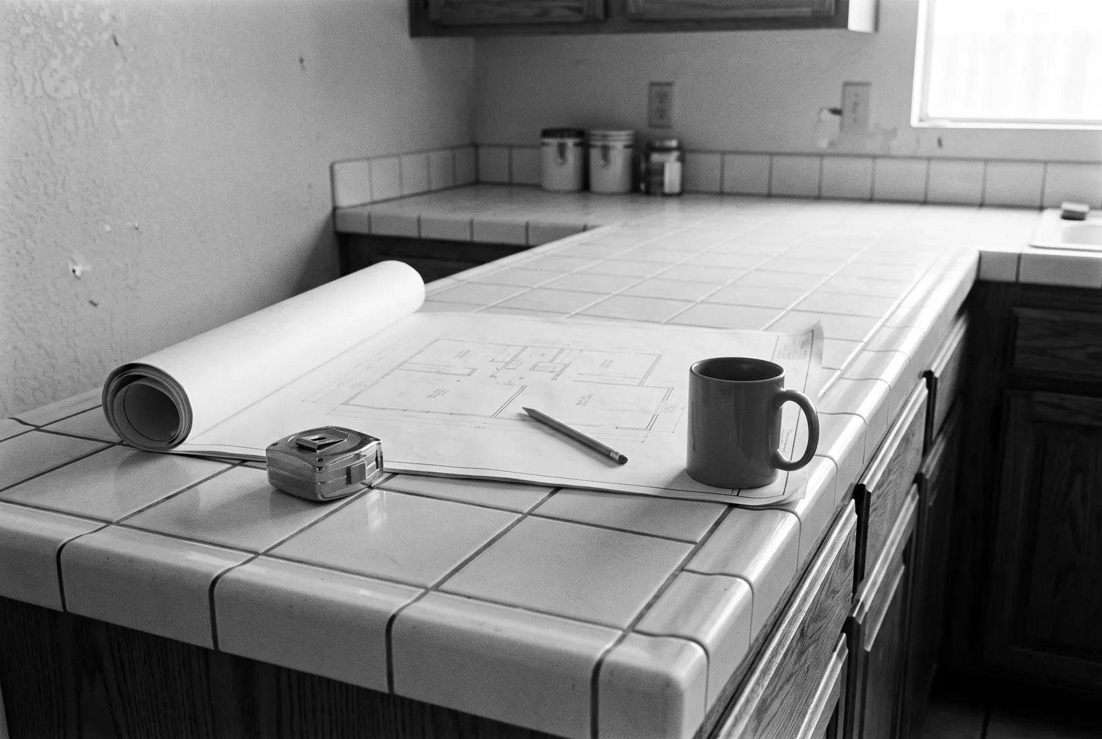 Render of a floor plan drawing half unrolled on an old tiled kitchen counter, with a pencil and a mug on it and a tape measure beside it.