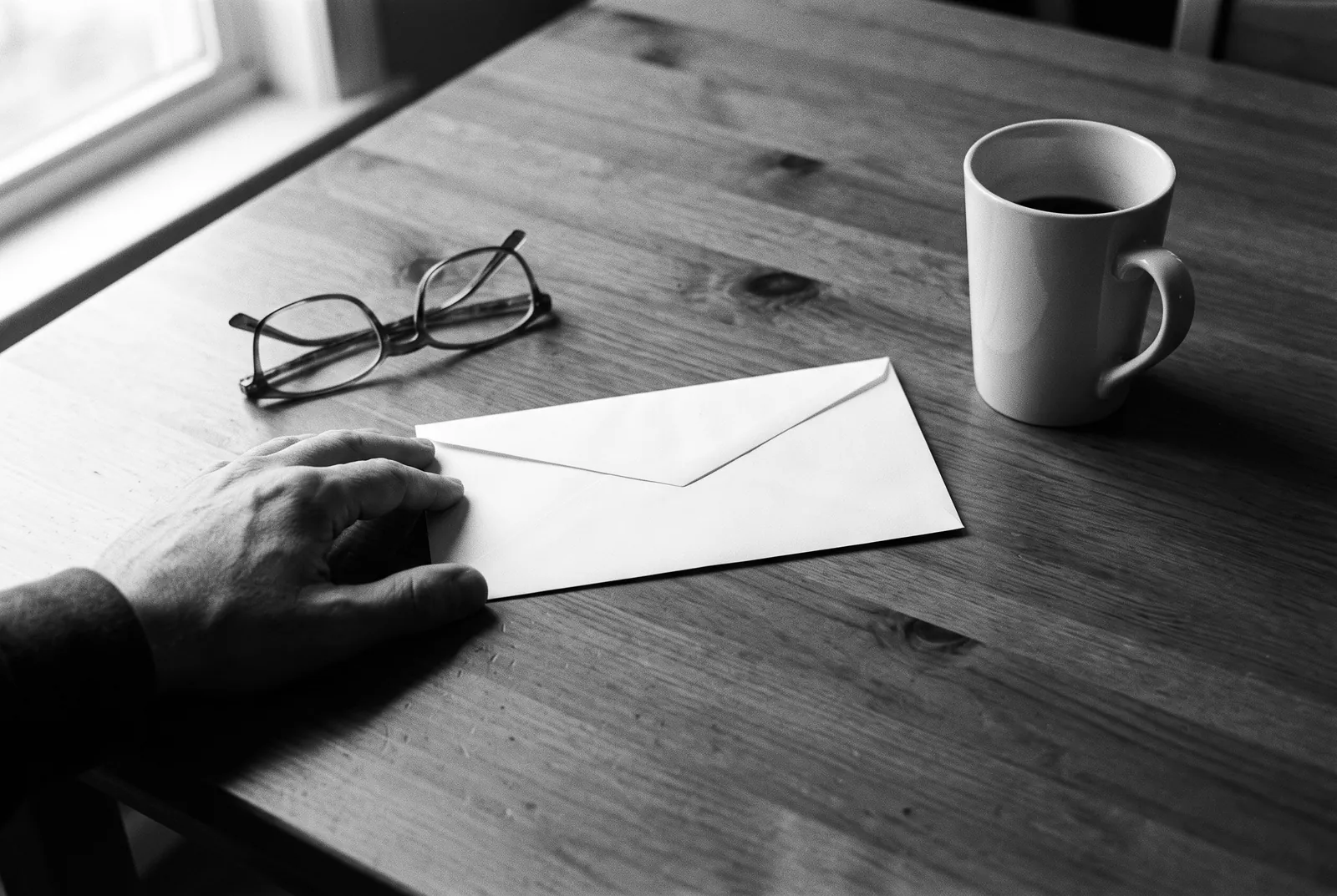 Render of a hand resting beside a sealed blank envelope on a wooden table, with reading glasses and a coffee mug.