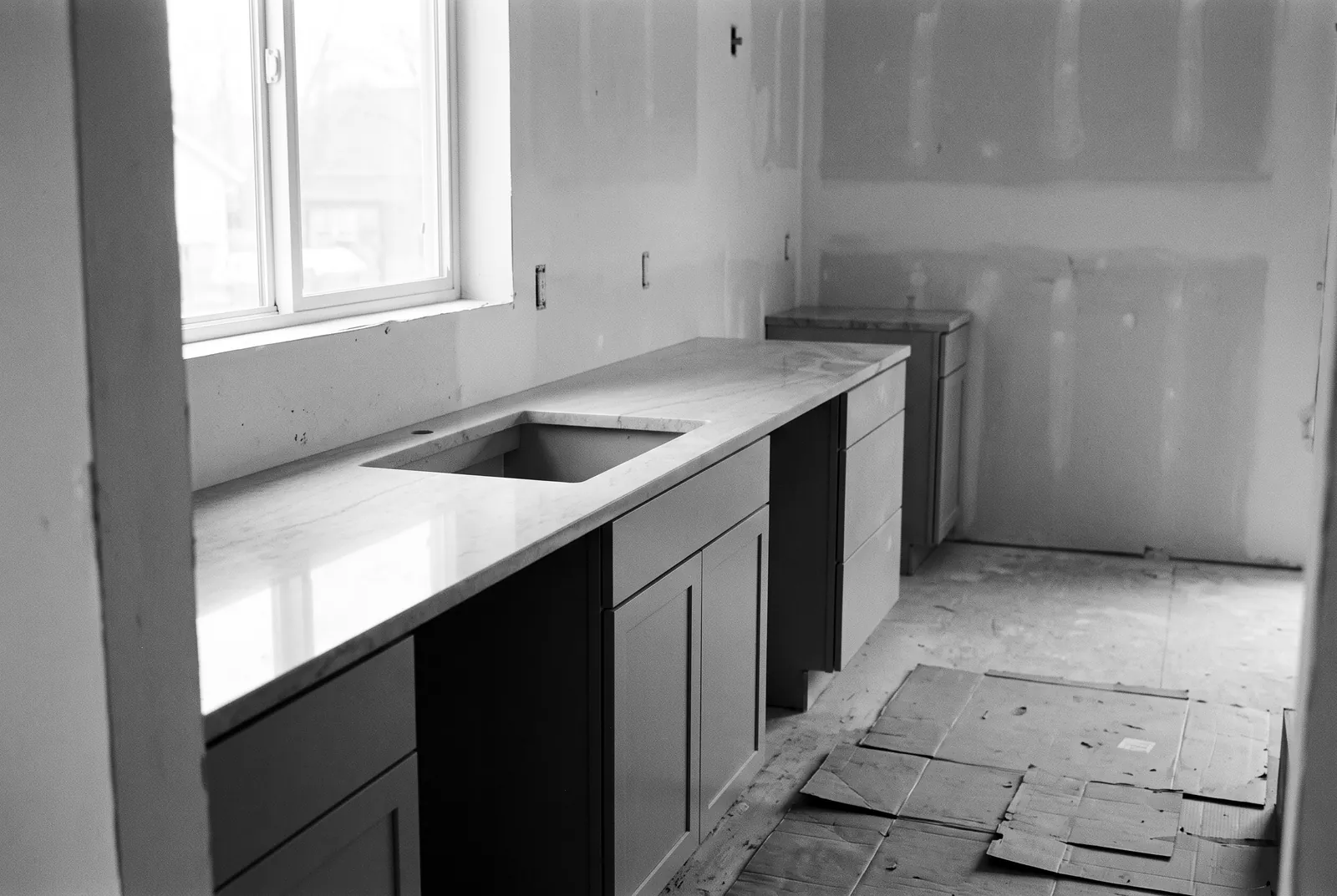 Render of new base cabinets along a wall with a stone top set on them and a sink cutout, in a room with bare drywall and cardboard on the floor.