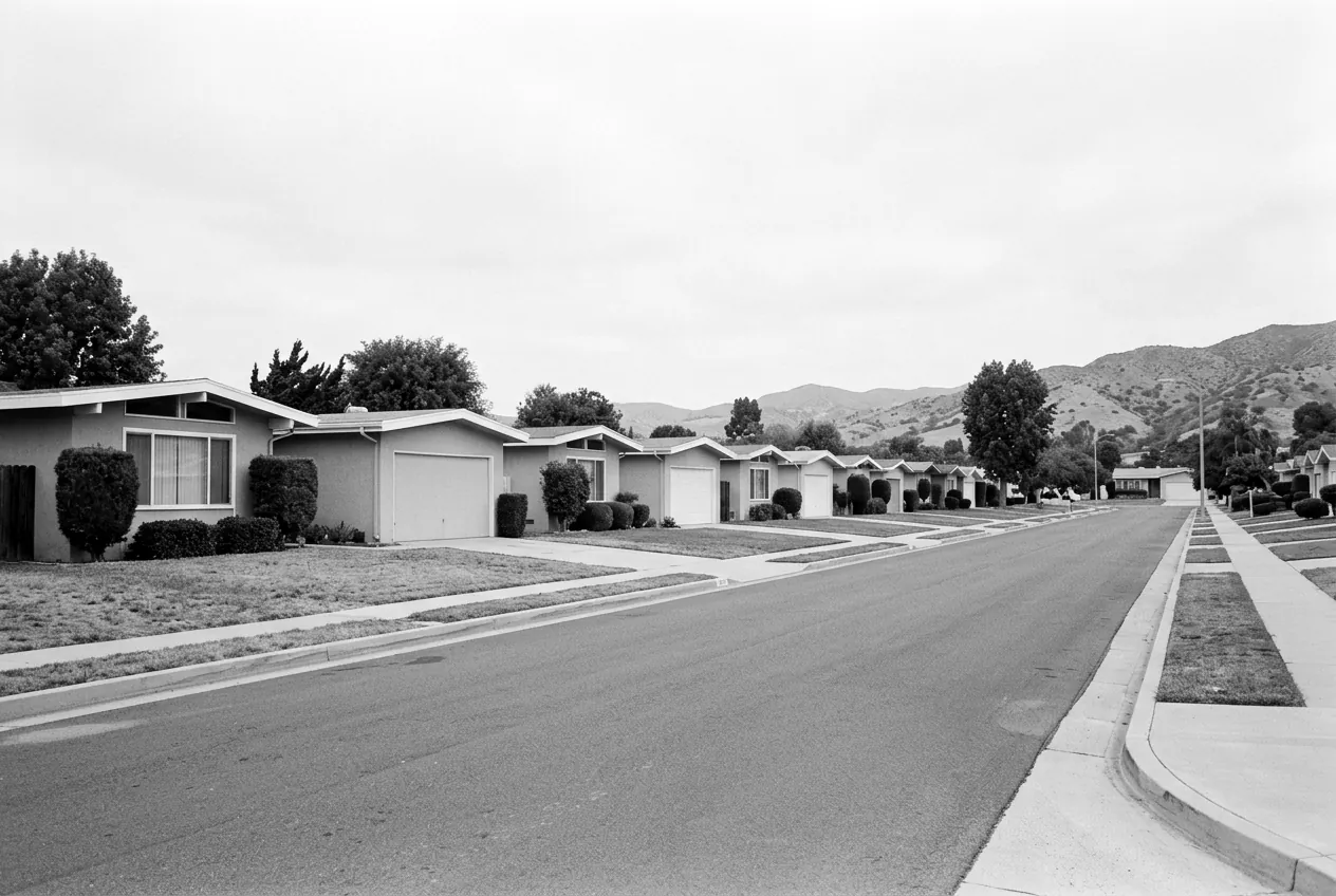 Render of a quiet street of single story tract houses from the 1960s, with low dry hills at the end of the block.