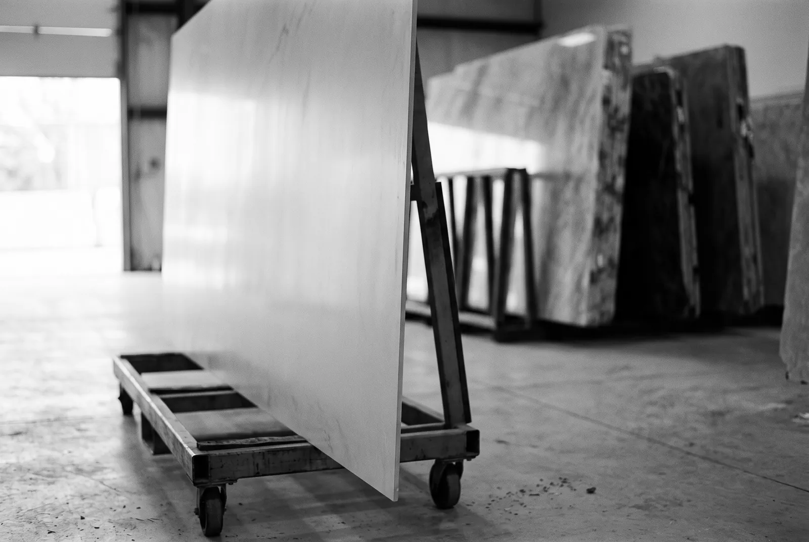 Render of a thin porcelain panel standing on a steel cart in a warehouse, with thicker stone slabs in a rack behind it.