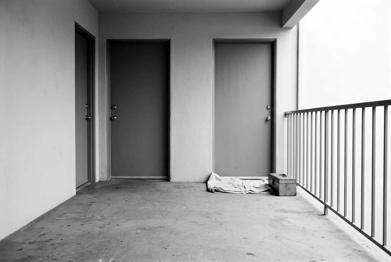 Render of two plain front doors a few feet apart on a shared concrete landing with a painted steel railing, a folded drop cloth and a toolbox by one door.