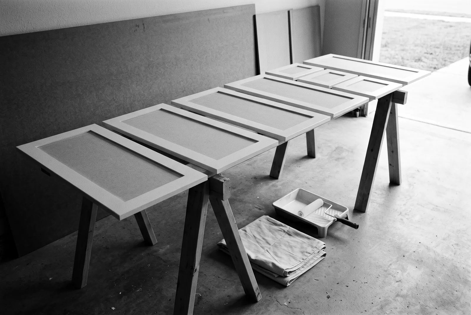 Render of unfinished shaker cabinet doors and drawer fronts laid across sawhorses in a garage, with a paint tray, a roller and a drop cloth on the floor.