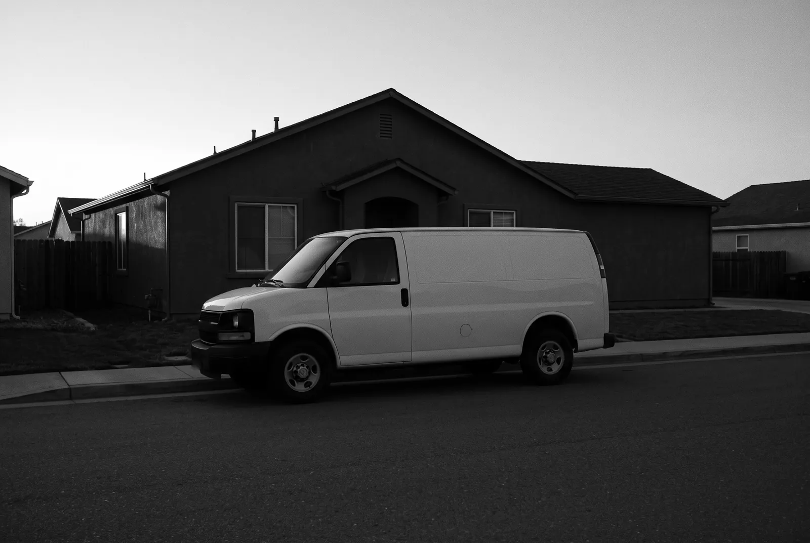 Render of a plain white work van parked at the curb in front of a dark stucco house before sunrise.