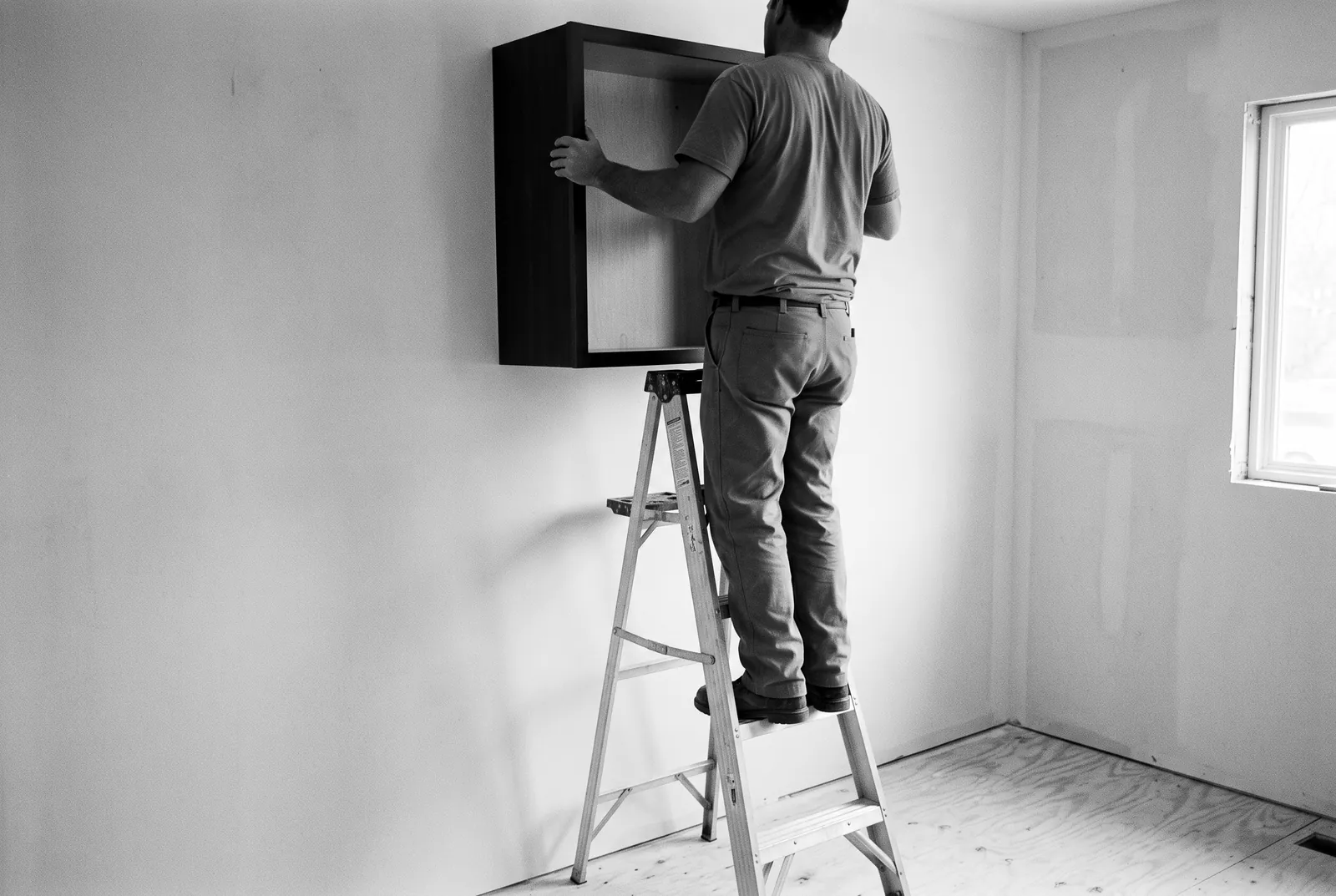 Render of a worker seen from behind on a step ladder, lifting an empty upper cabinet box into place on a bare wall.